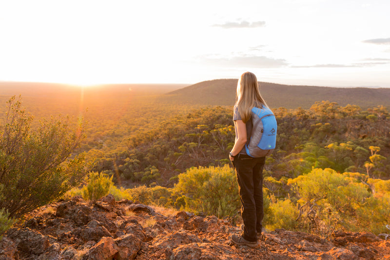 Female hiker with a blue Sea to Summit ultra-sil backpack standing on a rocky outcrop, watching the sunset over a forested valley