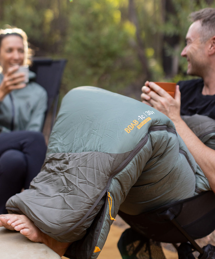 A man sitting in a camping chair, wearing the Boab sleeping bag. He is holding a cup and is chatting to a woman. 
