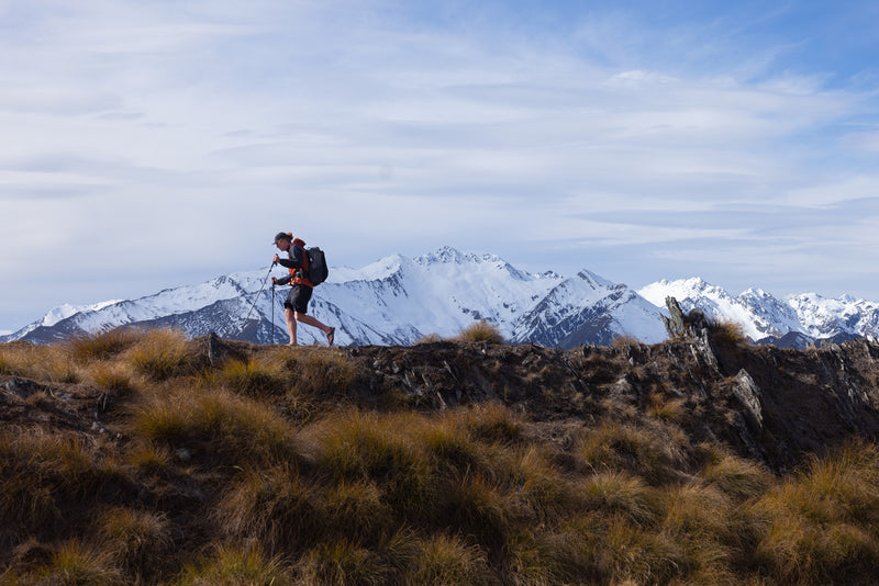 Hiker with backpack and trekking poles walking along a grassy alpine ridge with snow-covered mountains in the background