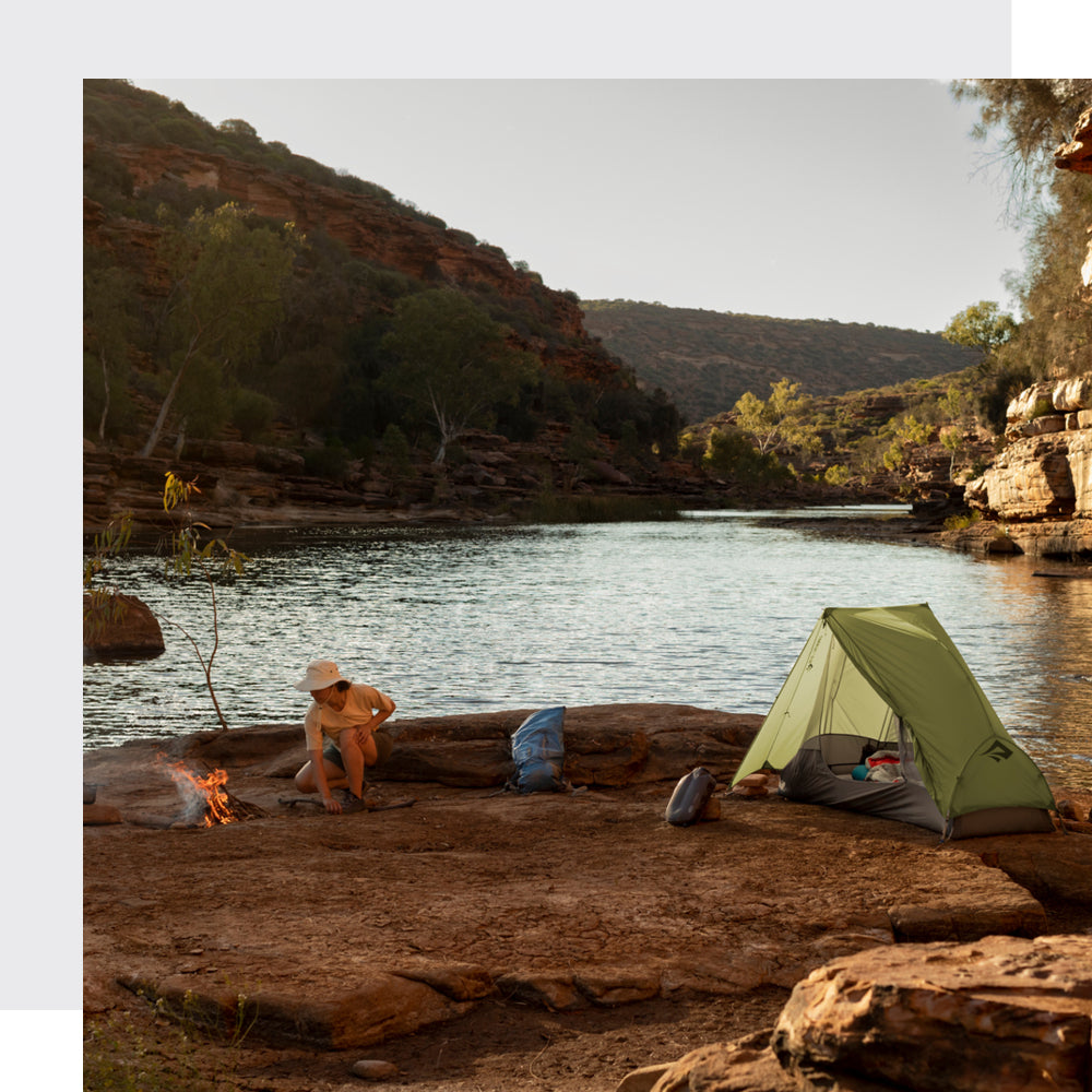 Person crouching by a small campfire near a river in a rocky canyon landscape, with a green tent pitched nearby. Camping gear is scattered around the site under the late afternoon sun.