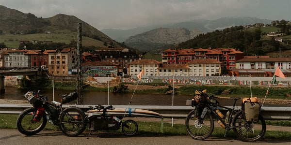 Two fully loaded bikepacking bicycles with gear and a surfboard trailer parked beside a river, with a Spanish town and mountains in the background