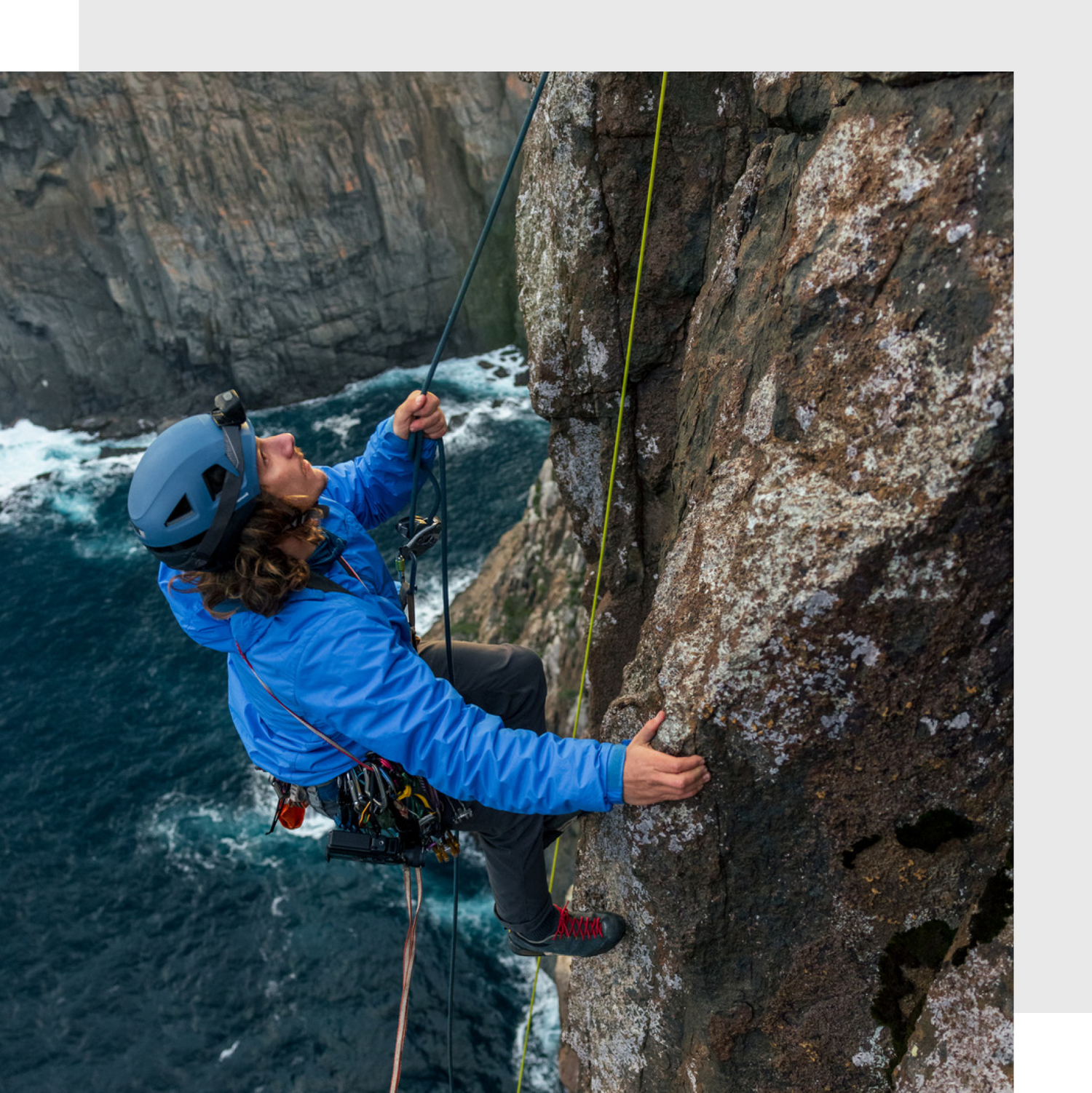 Rock climber in a blue jacket and helmet ascending a coastal cliff face with safety ropes and climbing gear, overlooking the ocean below.