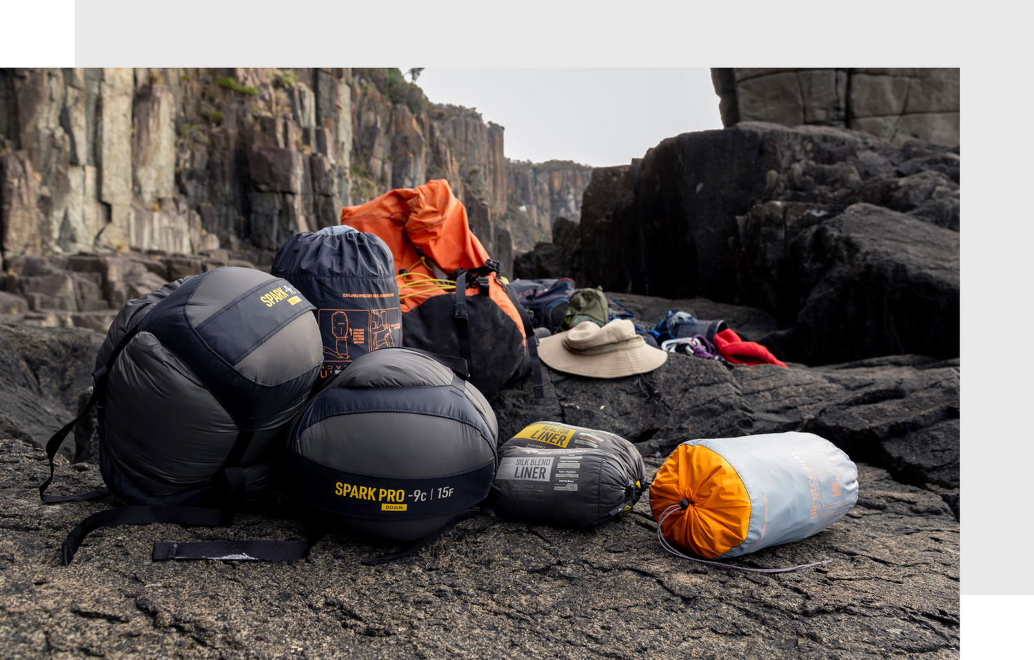 Close-up of compact camping gear, including sleeping bags labeled 'Spark Pro -9°C | 15°F', a silk blend liner, and a packed sleeping mat, laid out on rocky terrain with cliffs in the background. Additional gear, a backpack, and a sunhat are scattered nearby.
