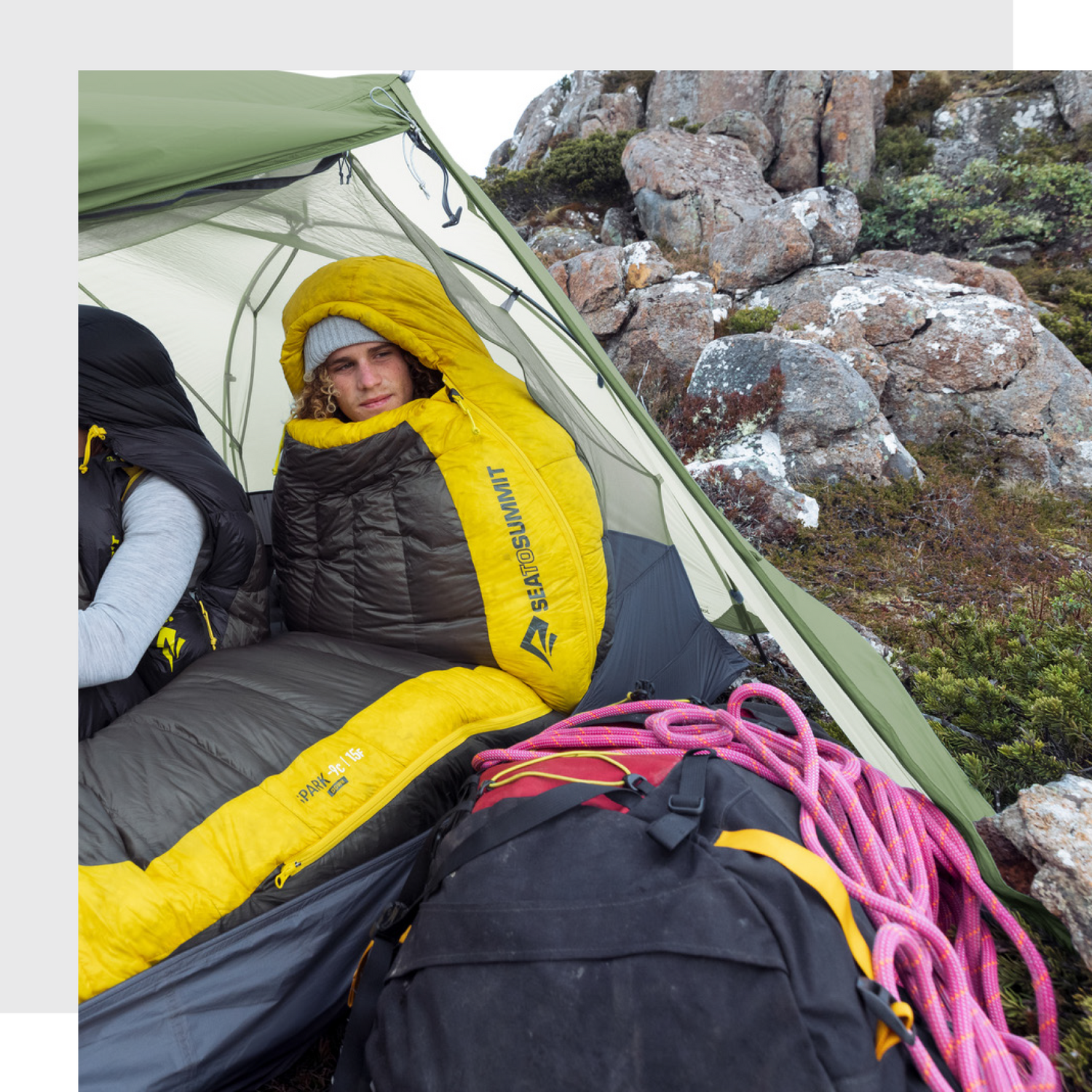 Person inside a Sea to Summit Spark sleeping bag, resting in a tent pitched on a rocky mountain terrain, with climbing rope and a backpack visible in the foreground.